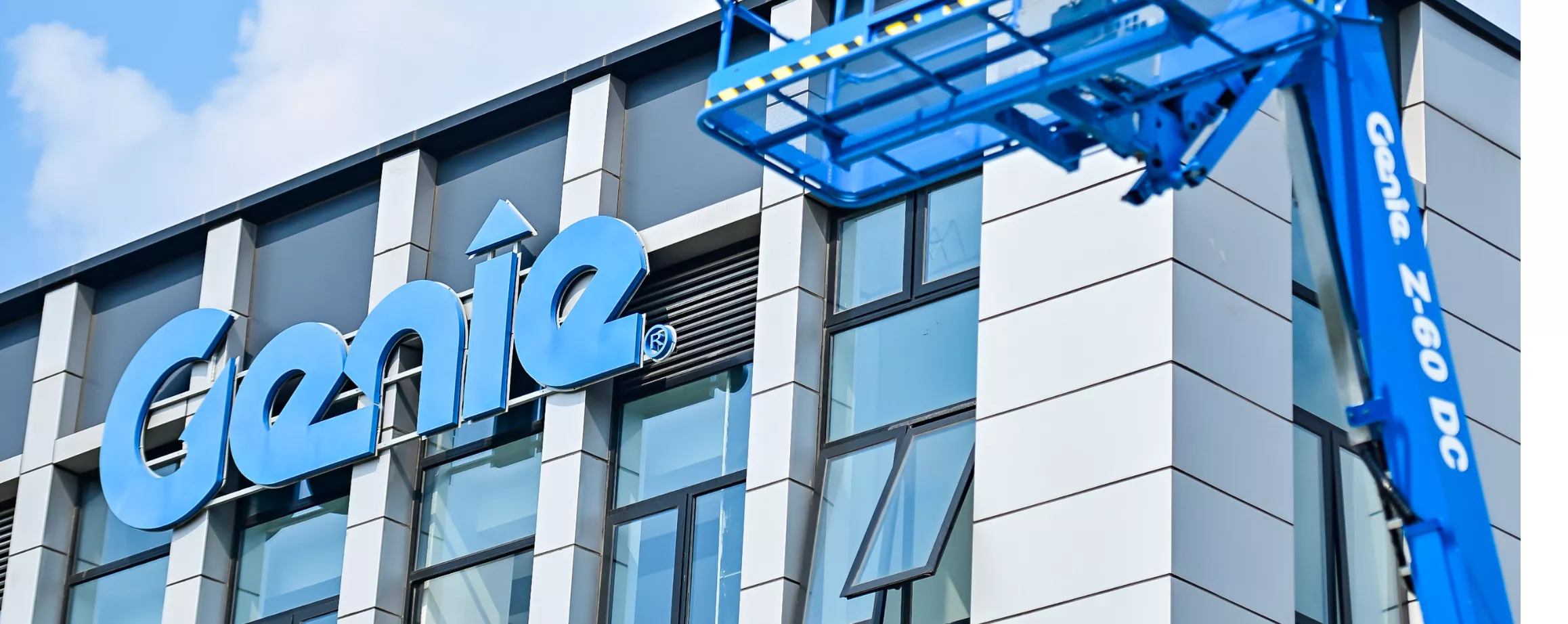 A blue Genie Z-60 DC aerial lift is positioned near a contemporary glass and metal building. The Genie logo is prominently displayed on the building facade. The lift's platform is extended upward, showcasing its industrial design and functionality.